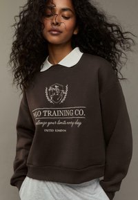 Felpa marrone con colletto bianco, con logo ricamato e testo: "POLO TRAINING CO. sfida i tuoi limiti ogni giorno, REGNO UNITO."