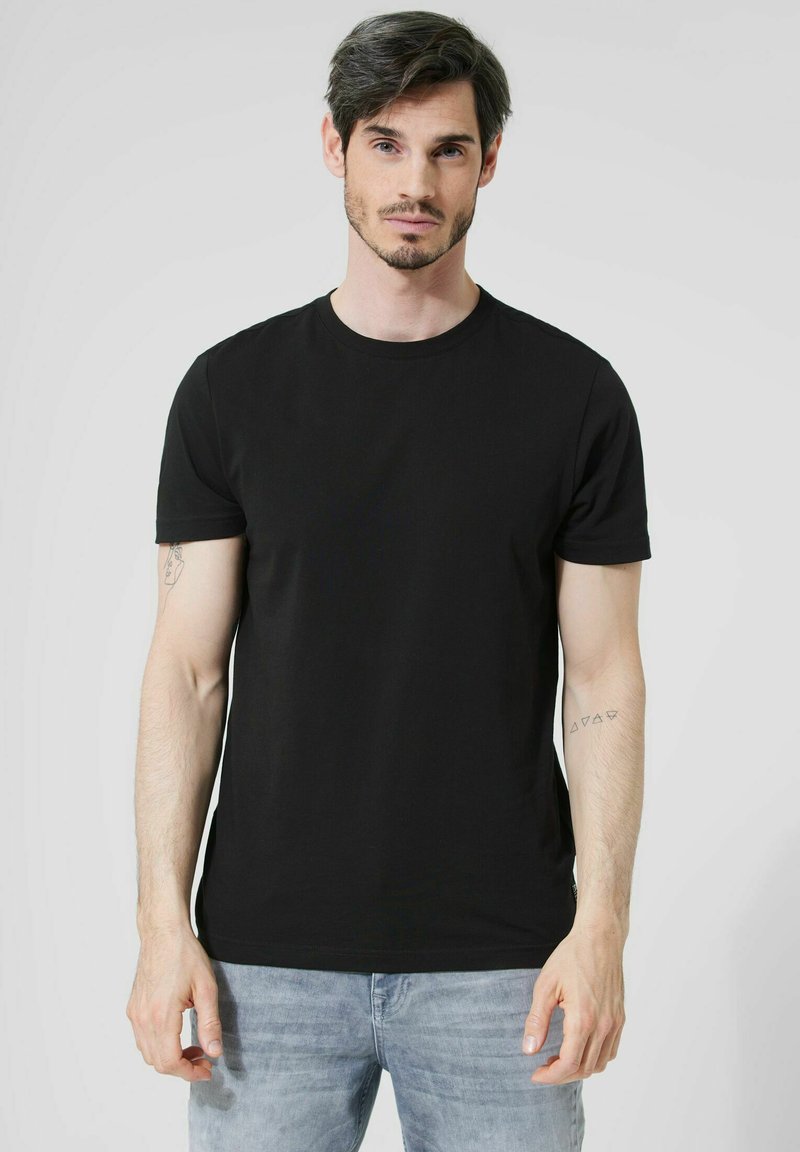 Černé tričko s krátkým rukávem vyrobené z bavlny s kulatým výstřihem. Model ukazuje tetování na levém rameni. Jednobarevné bez vzorů.