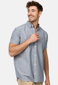 Koszula z krótkim rękawem w jasnoniebieskim kolorze wykonana z chambray, z kołnierzykiem w kształcie szpica oraz małym granatowym logo na lewej kieszeni piersiowej.