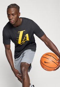 Musta T-paita, jossa keltainen "LA"-kuvio, harmaat shortsit, oranssi koripallo kädessä, keskittynyt asento, sileä kangas, urheiluvaatteet.
