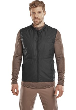 Gilet isolant noir avec un col haut et une fermeture éclair sur le devant, porté sur un haut à manches longues marron ; texture lisse ; et design simple.