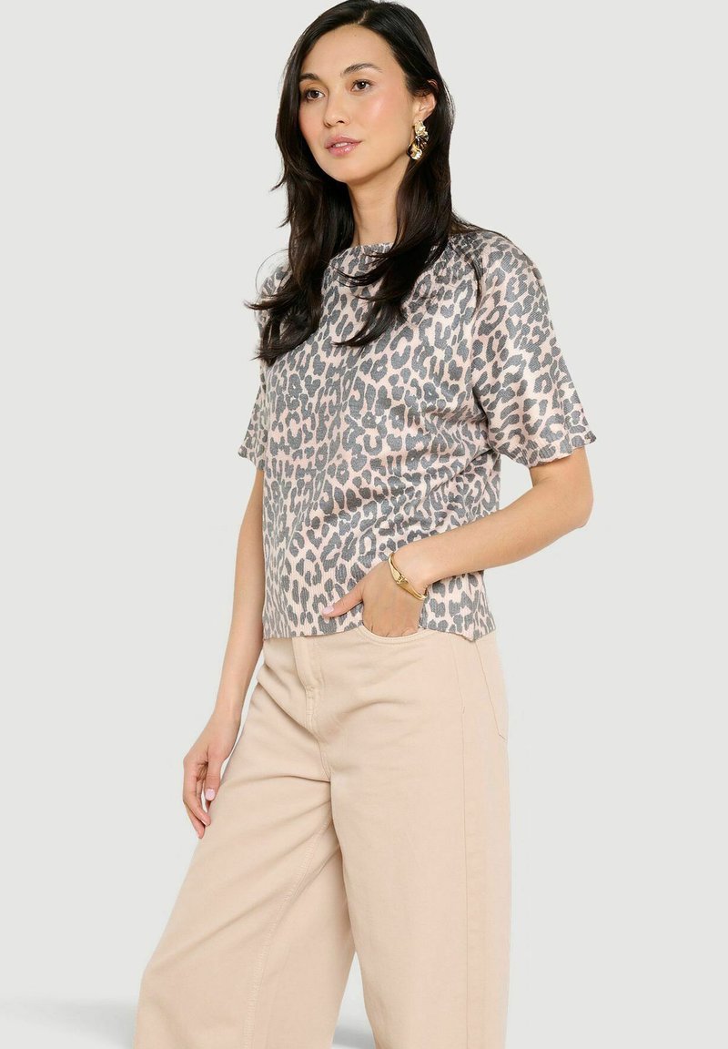 Mujer con cabello largo y oscuro, vestida con una blusa de manga corta de estampado de leopardo, pantalones anchos beige, pulsera y pendientes dorados, de pie con una mano en el bolsillo.