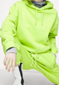 Limegrön Nike-hoodie och matchande sweatpants med dragsnoddar. Modellen bär en grå undertröja och tydliga svarta och vita stövlar.