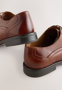 Paire de chaussures habillées en cuir marron poli avec semelles en caoutchouc noir, coutures visibles et talon arrondi sur fond clair.