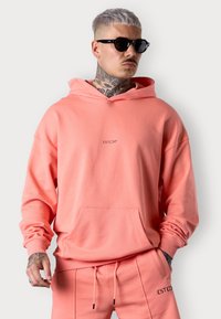Sweat à capuche rose corail avec une poche avant, des poignets côtelés et une capuche avec cordon de serrage. Présenté avec un pantalon de jogging assorti et des lunettes de soleil noires.