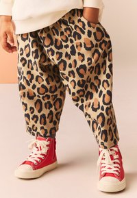 Pantalons beige à motif léopard, avec une coupe ample, présentant des taches noires et beiges. Associés à des baskets montantes rouges avec des lacets blancs.