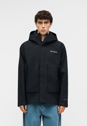 Młody mężczyzna z kręconymi włosami, ubrany w czarną kurtkę z kapturem marki Columbia i niebieskie dżinsy, stoi na tle jasnego, jednolitego tła.