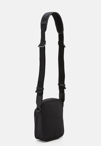 Sac bandoulière noir en matériau lisse de forme rectangulaire. Doté d'une sangle ajustable avec un logo en relief et d'une fermeture éclair.