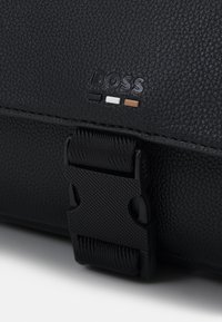 Borsa in pelle nera con superficie texturizzata, una chiusura a fibbia evidente e un logo con accenti bianchi, neri e marroni.