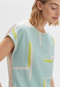 Blouse bleu clair en tissu doux avec des motifs abstraits en blanc et vert lime. Encolure ronde et manches larges.