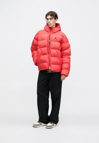 Chaqueta de abrigo roja con cuello alto y cremallera, con secciones acolchadas y un logo negro. Combinada con pantalones negros y zapatillas.