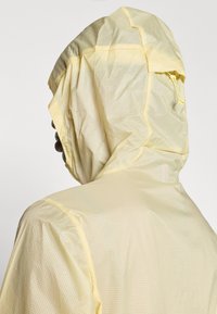 Ανοιχτό κίτρινο αδιάβροχο μπουφάν με κουκούλα, με υφή υφάσματος και αντίθετη κίτρινη ραφή, διαθέτον τας τσέπη με φερμουάρ στην πλάτη.