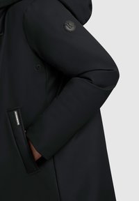 Zwarte buitenkleding met een gladde, matte afwerking, een label aan de zijkant en een ronde logo patch op de mouw, met een ontspannen pasvorm ontwerp.