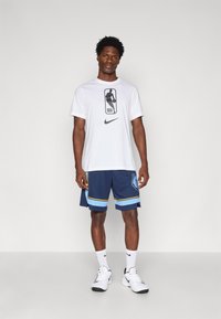 T-shirt branca da Nike com o logótipo da NBA, combinada com calções azul-marinho com um gráfico de urso e detalhes em amarelo e azul, complementada por ténis brancos.