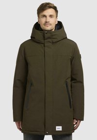 Olive green parka jacket with a large hood, front zipper and snap closure, two side pockets with zippers, and a logo patch at the hem.
