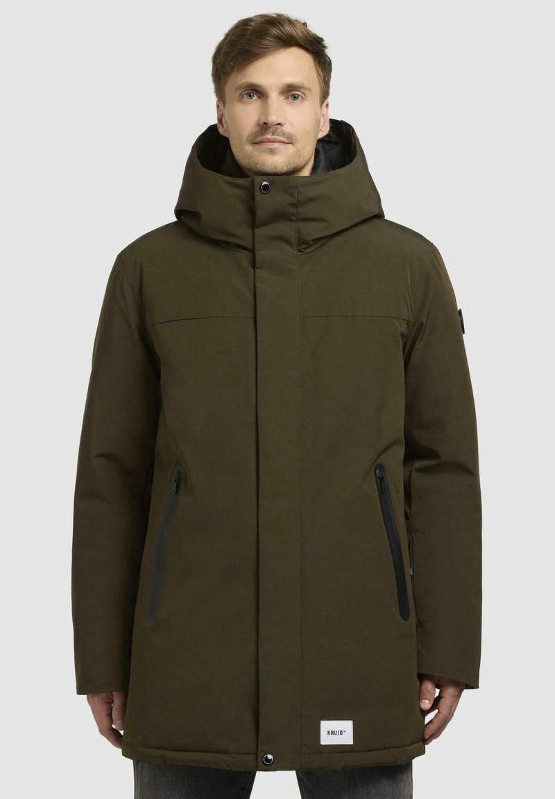 Olive green parka jacket with a large hood, front zipper and snap closure, two side pockets with zippers, and a logo patch at the hem.