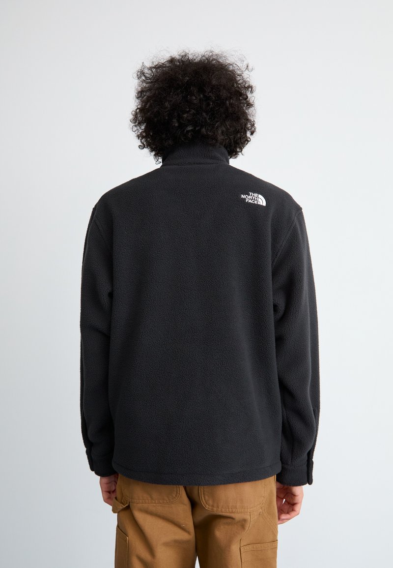 Veste en polaire noire avec col montant et manches longues, ornée d'un petit logo blanc dans le haut du dos, portée avec un pantalon cargo beige.