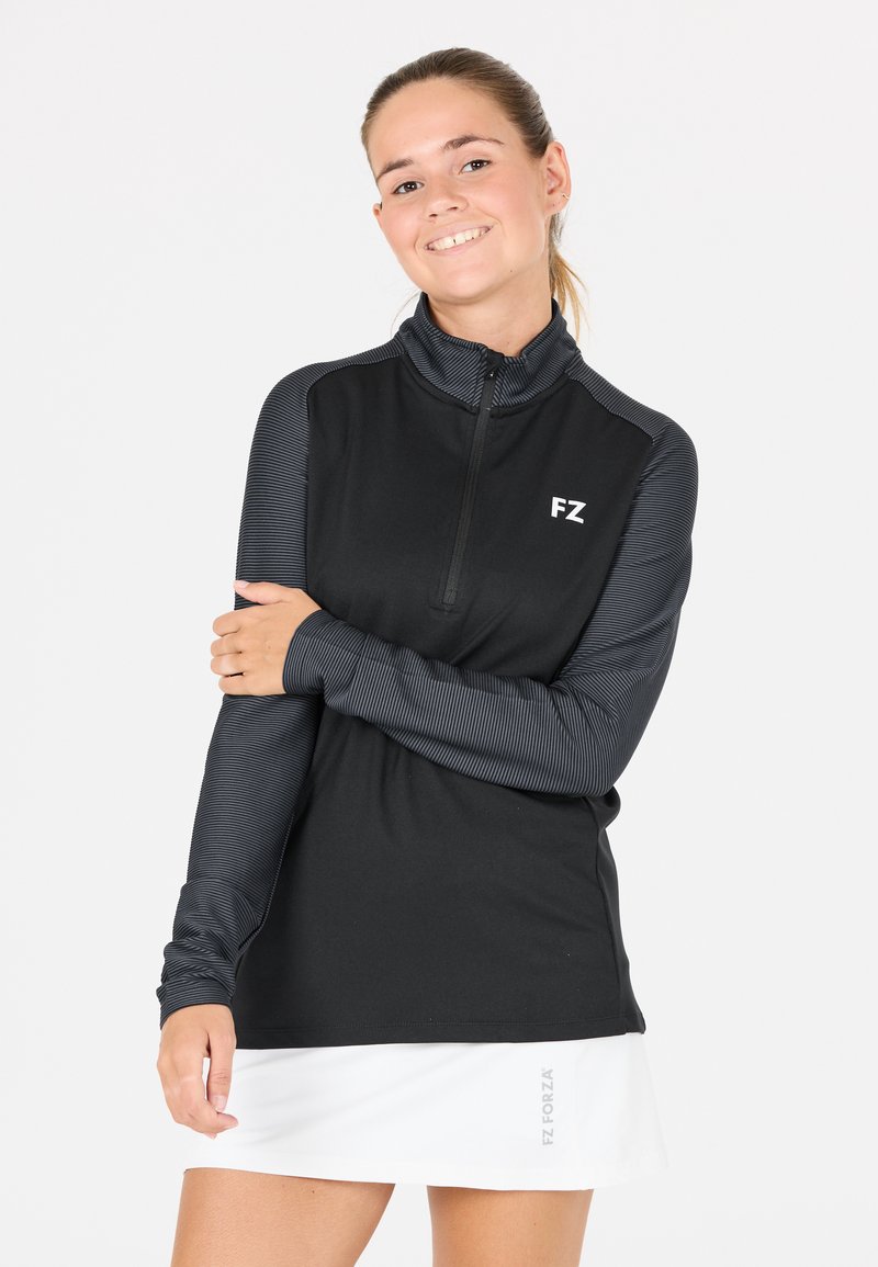 Ung kvinna i en svart och grå långärmad sporttröja och en vit kjol, leende med armarna korsade över bröstet.