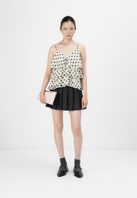 Femme dans une jupe plissée noire, un top à volants blanc à pois, des chaussettes noires jusqu'aux chevilles, des chaussures noires, tenant une pochette rose sur fond blanc.