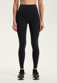 Czarne legginsy slim fit noszone z czarno-białymi butami sportowymi, pokazane od talii do stóp na jednolitym jasnym tle.