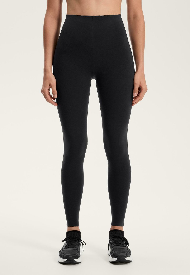 Czarne legginsy slim fit noszone z czarno-białymi butami sportowymi, pokazane od talii do stóp na jednolitym jasnym tle.