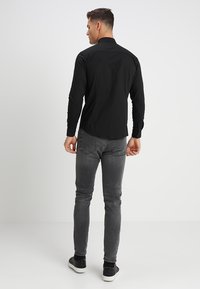 Schwarzes Langarmshirt aus Baumwolle mit klassischem Kragen und Rückenpasse, kombiniert mit dunkelgrauen Slim-Fit-Jeans und schwarzen Sneakers.