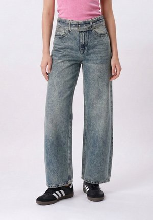 Osoba w wysokich, szerokich, wyblakłych niebieskich dżinsach z wysokim stanem i pasem w tym samym kolorze, różowej bluzce oraz czarnych trampkach z białymi paskami.