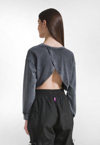 Sudadera gris recortada con un corte único en la espalda, mangas largas y un ajuste cómodo. Combinada con pantalones negros con cinturilla elástica y una etiqueta rosa.