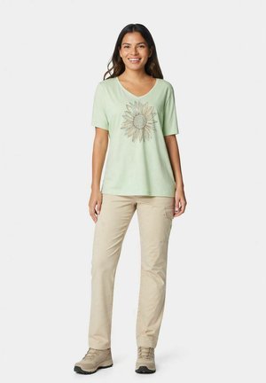 Donna sorridente con maglietta verde chiaro con grande stampa di girasole, pantaloni cargo beige e scarpe da trekking beige, in piedi su sfondo bianco.