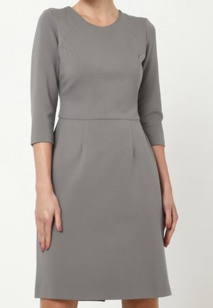 Femme portant une robe grise mi-longue aux manches trois-quarts et taille cintrée sur un fond blanc uni.