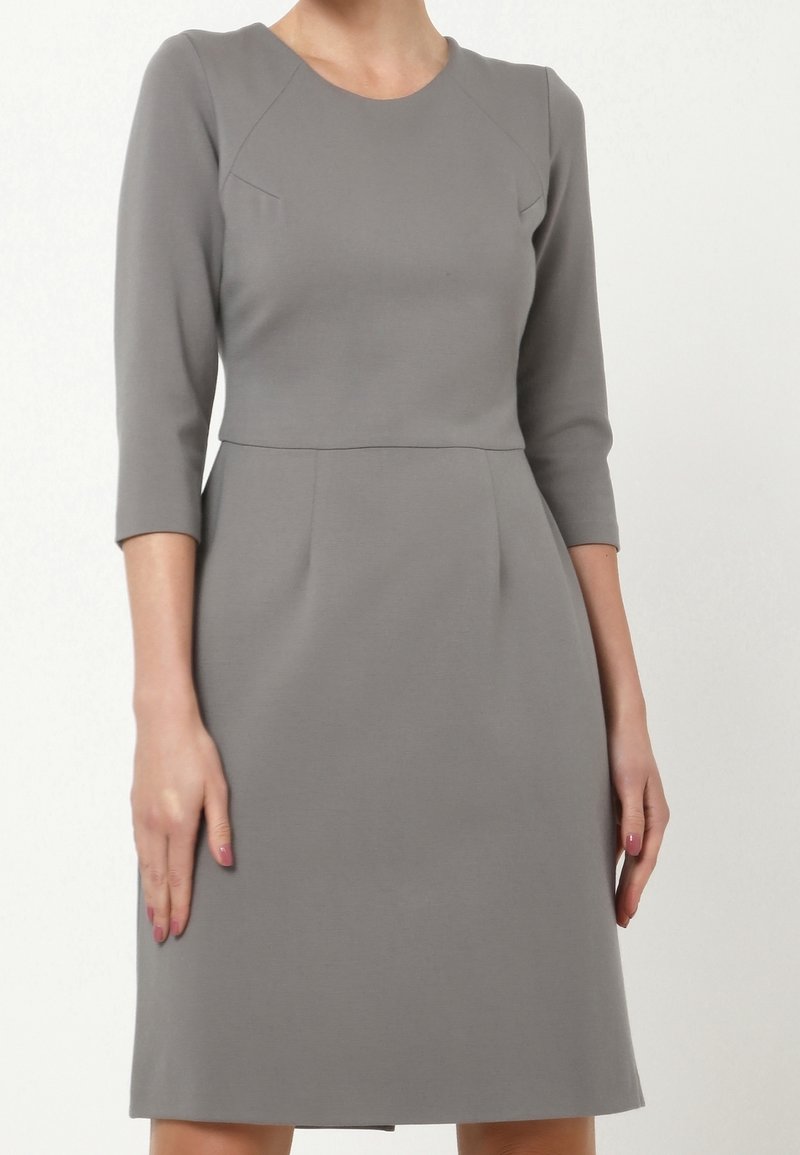 Femme portant une robe grise mi-longue aux manches trois-quarts et taille cintrée sur un fond blanc uni.