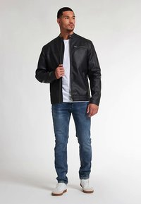 Giacca di pelle nera con colletto alla coreana, chiusura frontale con zip e tasche laterali. Indossata sopra una T-shirt bianca e abbinata a jeans blu e sneaker bianche.