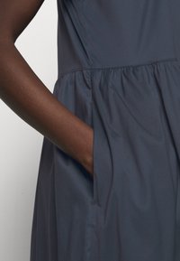 Robe bleu marine avec une surface texturée, présentant une taille froncée et une poche latérale, partiellement obscurcie par une main.