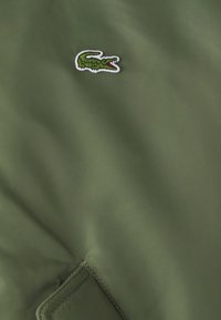 Olivově zelená bunda s hladkou texturou, na levé straně hrudi zdobená vyšívaným zeleným krokodýlím logem.