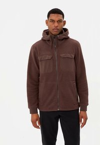 Bruine fleecejas met een ritssluiting aan de voorkant, twee borstzakken, een capuchon en een gestructureerde stof. Voorzien van een gladde zoom en zijzakken.