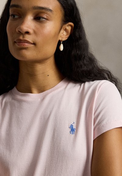 Γυναίκα με μακριά σκούρα μαλλιά που φοράει ανοιχτό ροζ πουκάμισο με μπλε κεντημένο λογότυπο και χρυσά σκουλαρίκια με λευκά κρεμαστά κοχύλια.