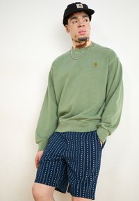Grønn bomullssweatshirt med ribbestrikkede mansjetter, sammen med marineblå shorts med et vertikalt hvitt prikkemønster. Svart caps med logo.
