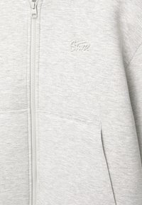 Sweat-shirt gris clair zippé avec poche frontale et logo brodé subtil sur le haut de la poitrine, montrant la texture du tissu et les détails des coutures.