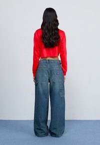 Rotes, gekürztes Langarmoberteil, kombiniert mit blauen Jeanshosen im weiten Bein, die mehrere Taschen und eine strukturierte Oberfläche aufweisen.