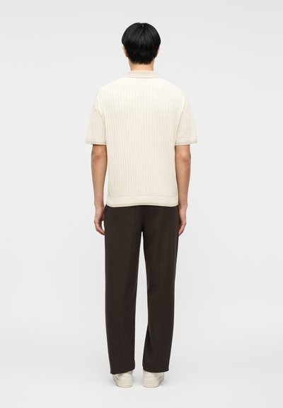 Homme debout de dos, portant un pull court à manches courtes en tricot crème, un pantalon marron foncé et des baskets blanches sur un fond uni.