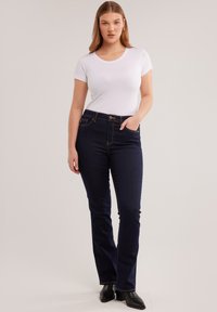 Žena odjevena u bijelu usku majicu, tamnoplave traperice i crne gležnjače, stoji s jednom rukom u džepu na jednobojnoj pozadini.