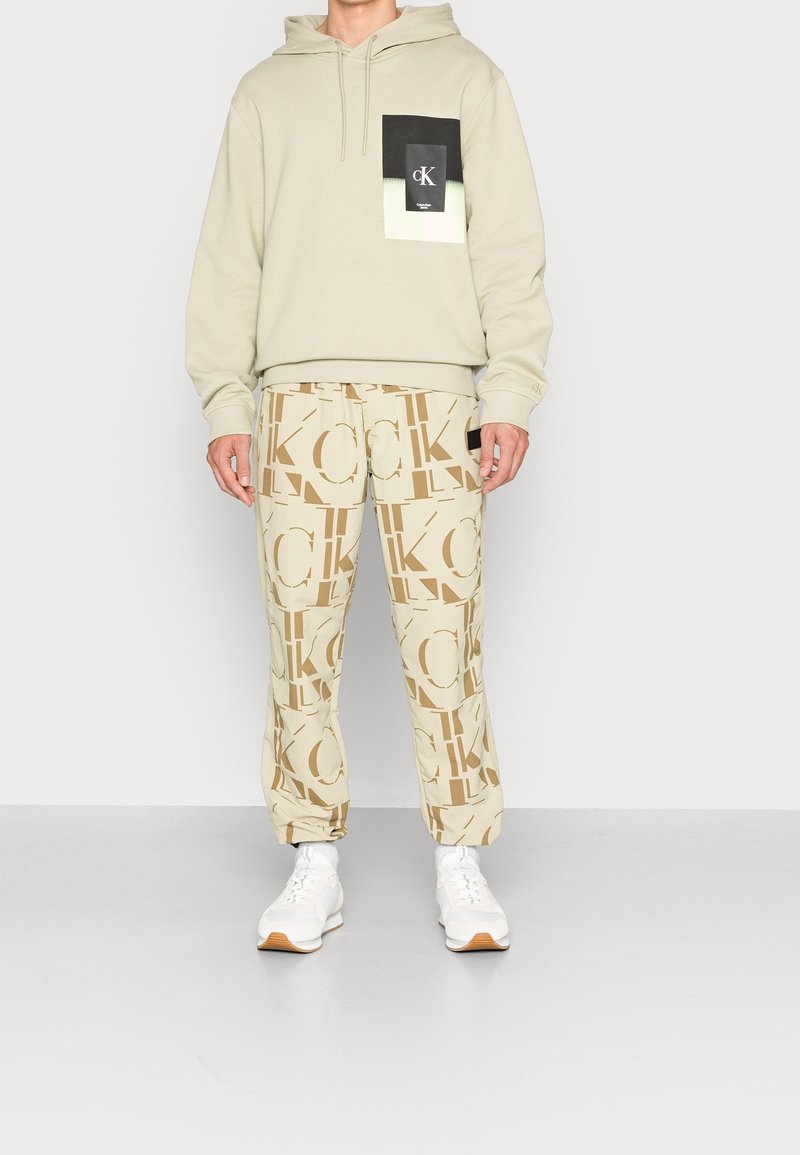 Homme portant un sweat à capuche beige avec un patch noir et blanc sur la poitrine, un pantalon beige avec de grandes lettres marron "CK" et des baskets blanches.