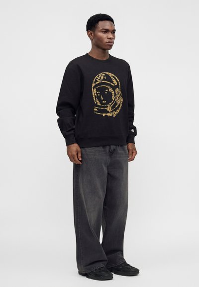 Άνδρας που φοράει μαύρο φούτερ με χρυσό γραφικό κράνος αστροναύτη, φαρδιά σκούρα γκρι τζιν και μαύρα αθλητικά, στέκεται με το πρόσωπο προς τα εμπρός.