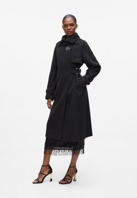 Black wool coat with a belted waist, high collar, and embroidered detail. Paired with a black sequined skirt and strappy black heels.