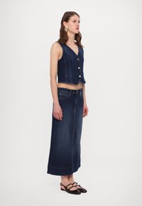 Denim vest with front buttons, paired with a long, denim skirt. Both in dark wash. Model wears black, strap shoes. Neutral background.