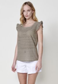 Top de punto caqui con escote cuadrado y detalles de hombros con volantes. Presenta recortes geométricos por toda la prenda para darle textura. Combinado con pantalones cortos blancos.