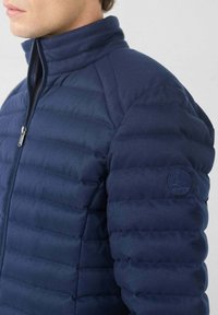 Hombre que lleva una chaqueta de plumón acolchada azul marino con cuello alto y cierre de cremallera, parche de logo visible en la manga izquierda.