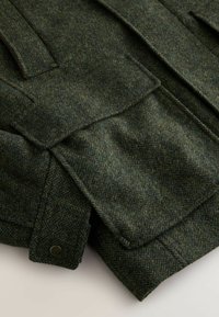Green herringbone wool fabric with a textured surface, featuring large front pockets and button detail. The design is structured and layered.