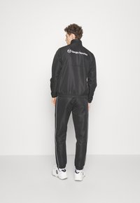 Fekete melegítőruha cipzáras dzsekivel, magas gallérral és Sergio Tacchini logóval. A nadrág fehér oldalsávokkal és rugós mandzsettákkal rendelkezik.