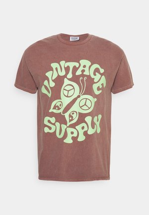 T-shirt en coton marron avec un grand motif graphique vert portant les mots "VINTAGE SUPPLY" et un motif de papillon. Manches courtes, col rond.